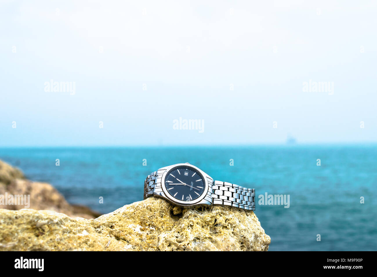 Eine exotische Edelstahl Silber watch gehalten auf der braunen Felsen am Strand und grünlichen Meer Wasser im Hintergrund bewölkter Tag Stockfoto