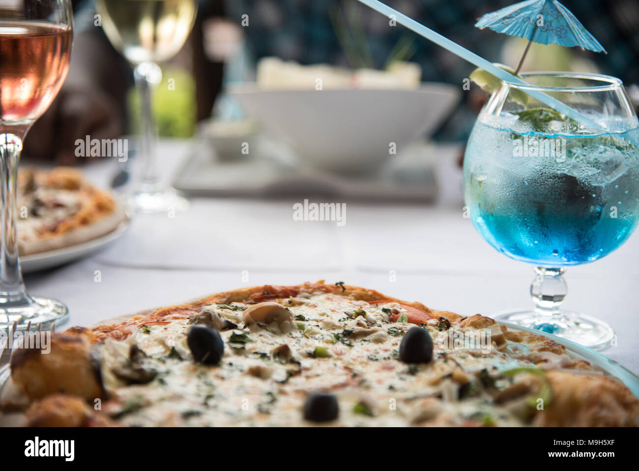 Foto von Restaurant Tisch mit zwei Pizzen und einen Salat. Auch ein helles Blau Cocktail und Glas rosa Wein. Stockfoto