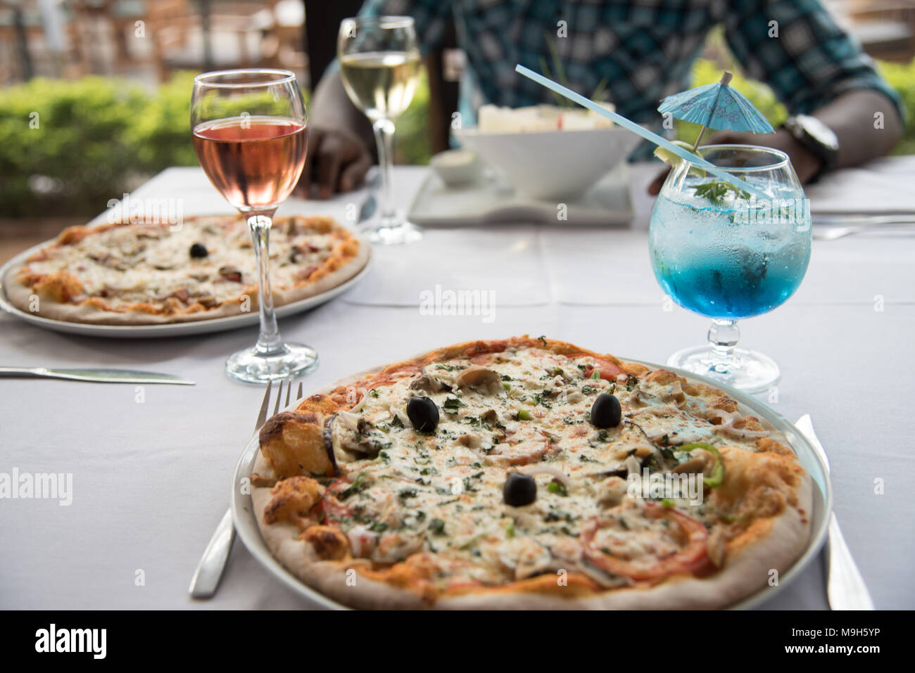 Foto von Restaurant Tisch mit zwei Pizzen und einen Salat. Auch ein helles Blau Cocktail und Glas rosa Wein. Ein dunkelhäutiger Mann sitzt im Hintergrund. Stockfoto