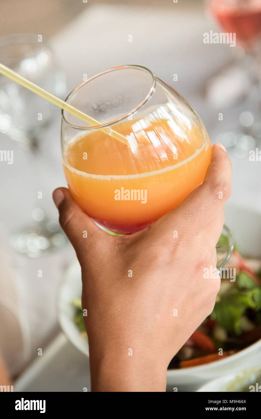 Die Hand eines weißen Mann hält ein helles Orange Saft/Cocktail. Essen und Gläser zu sehen in der Hintergrund verschwommen sein. Stockfoto