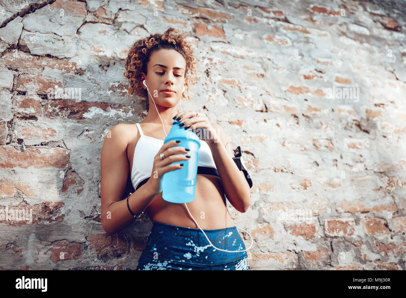 Portrait einer jungen muskulöse Frau mit Kopfhörer im Stehen an der Wand und trinken Wasser nach dem Joggen. Stockfoto