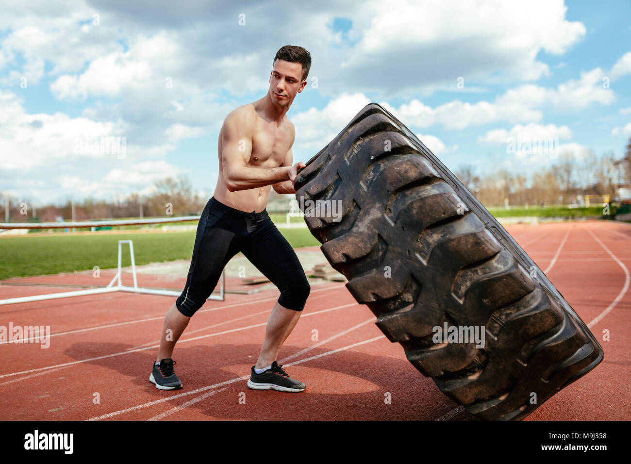 Junge muskulöse Mann hob eine riesige Reifen für Training Muskeln an der Tartanbahn auf dem Stadion. Stockfoto