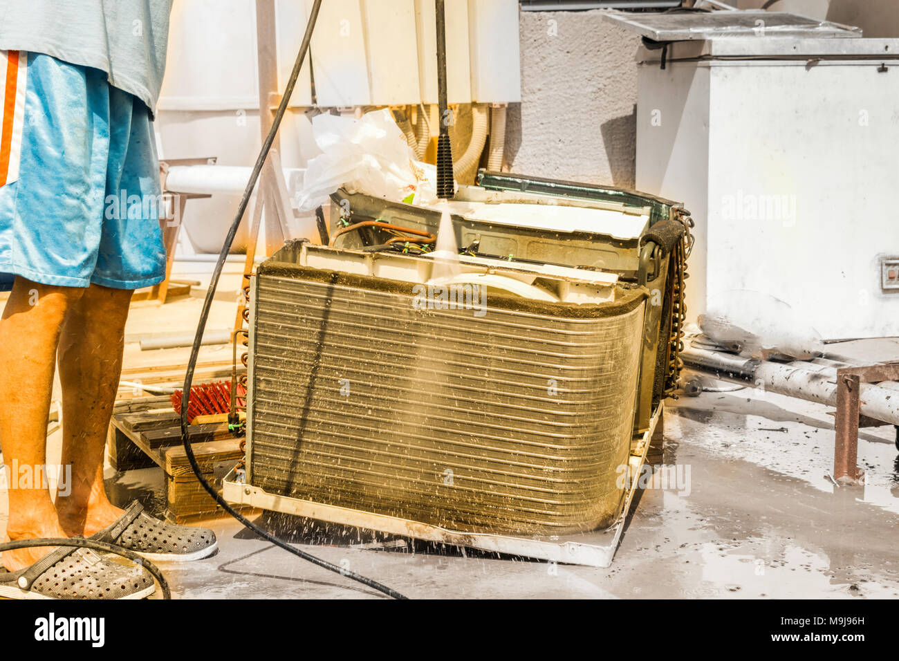 Einen professionellen Elektriker ist die Reinigung der Fenster Klimaanlage auf dem Dach eines Hauses mit seinem hohen Druck Wasser Pistole und trägt blaue Shorts Stockfoto