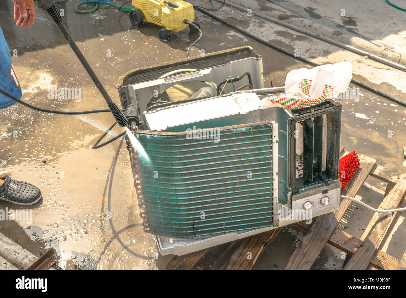 Einen professionellen Elektriker ist die Reinigung der Fenster Klimaanlage auf dem Dach eines Hauses mit seinem hohen Druck Wasser Pistole und trägt blaue Shorts Stockfoto