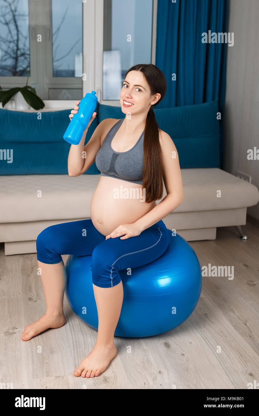 Glücklich schwanger Frau auf Fit Ball und Getränke Wasser, zu Hause zu sitzen. Die werdende Mutter. Schwangerschaft, Gymnastik für Schwangere, gesunden Lebensstil Konzept Stockfoto