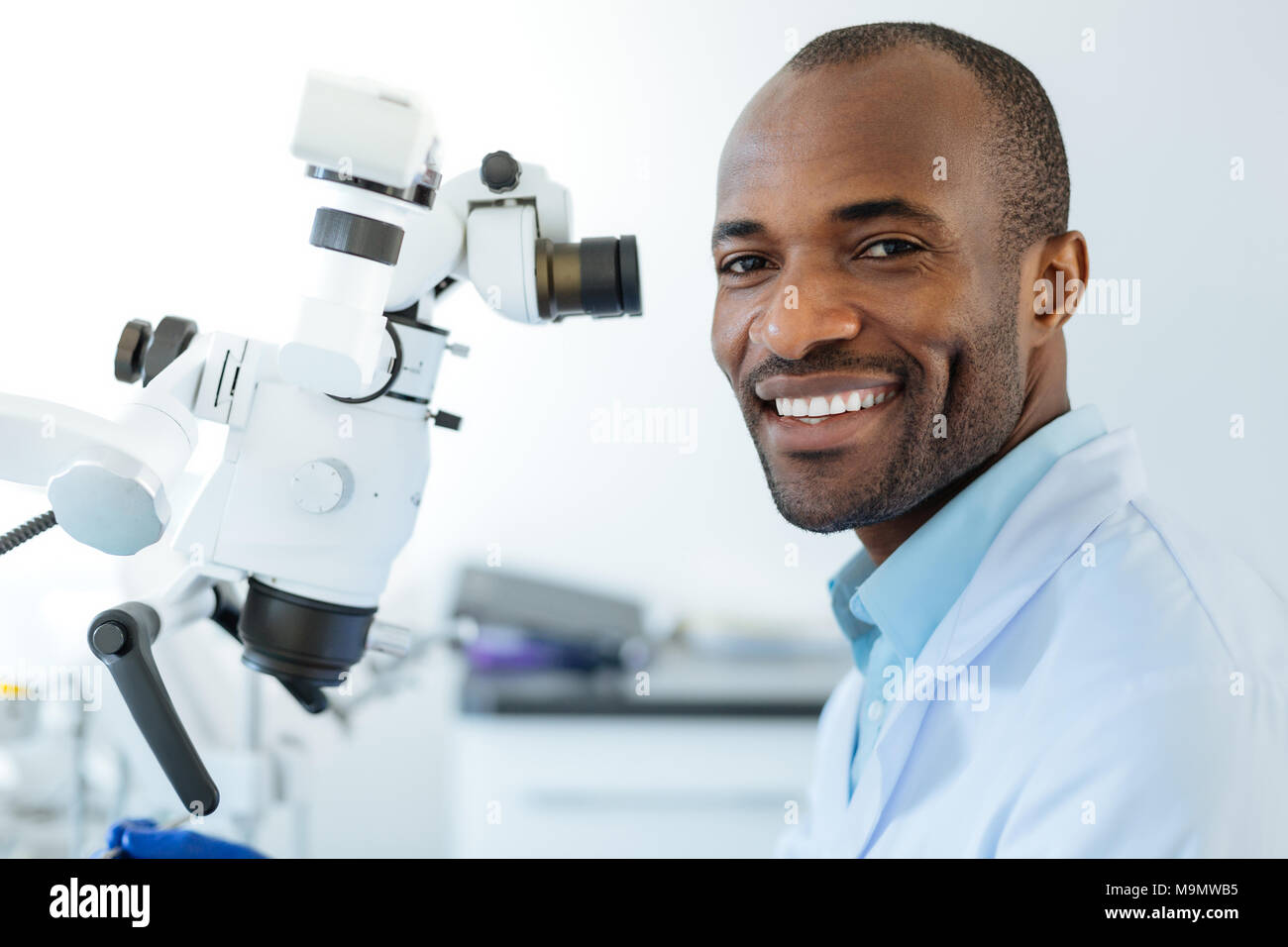 Glückliche Benutzer. Charmante upbeat Zahnarzt posieren für die Kamera und hell lächelnd, während Sie ein Mikroskop bei der Arbeit und die Durchführung von checkup Stockfoto
