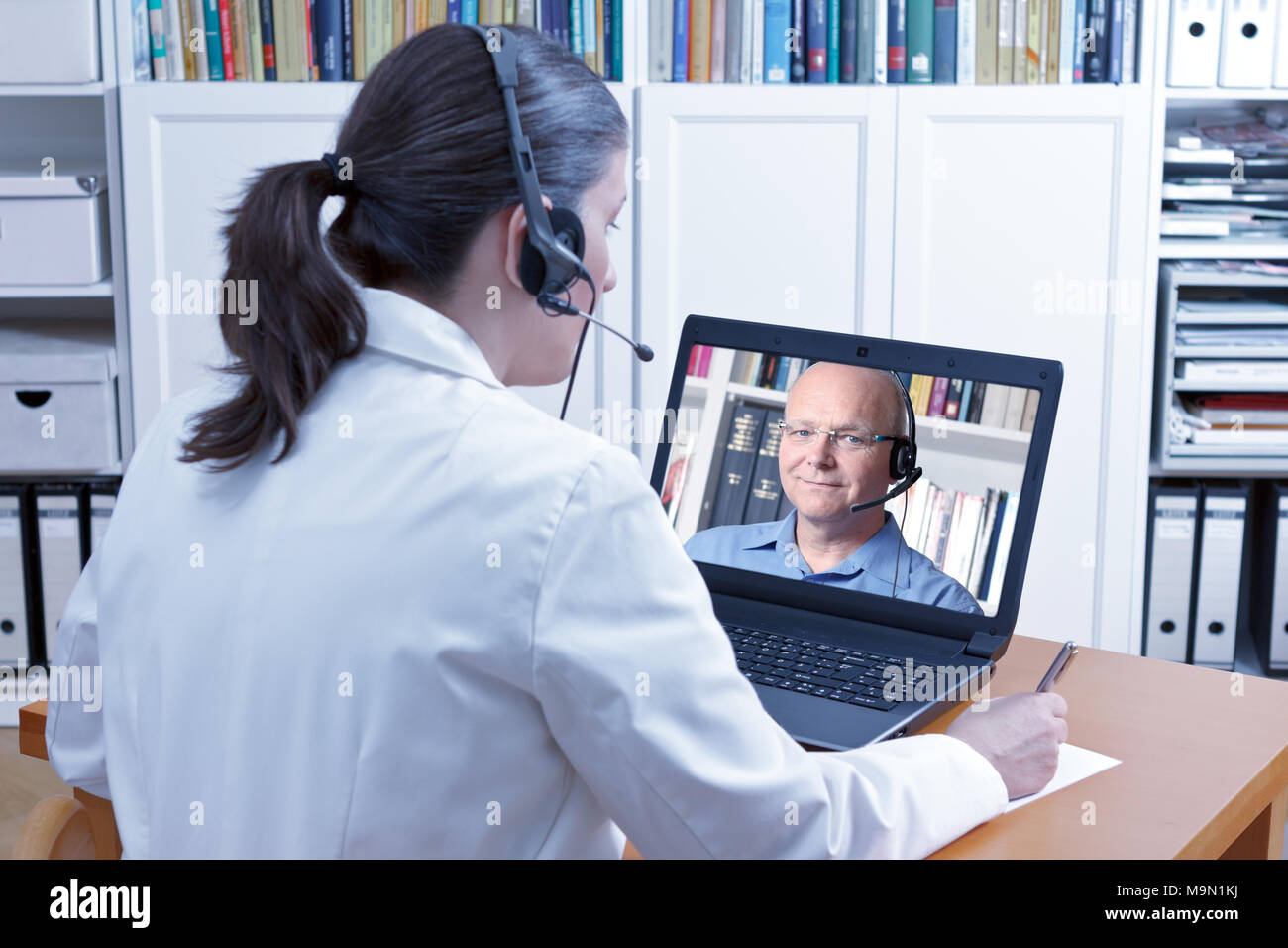 Arzt oder Apotheker sitzen in Ihrem Büro mit Headset und Laptop, sich Notizen während einer Videokonferenz mit einem männlichen Patienten, telehealth Konzept Stockfoto