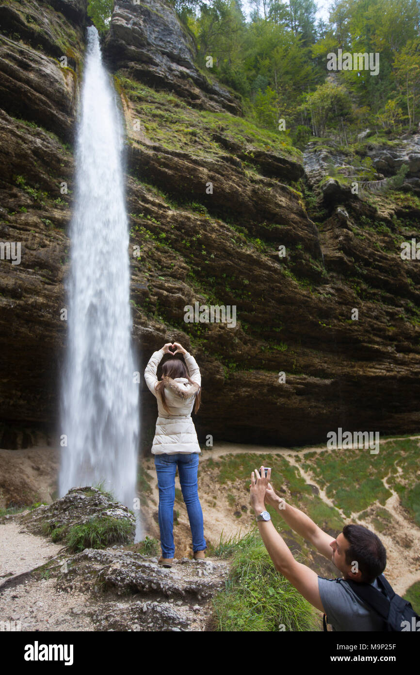 Slowenische Mädchen macht Herz Abbildung mit ihren Händen vor Wasserfall Pericnik in alpine Vrata Tal in der Nähe von Mojstrana im Triglav Nationalpark, während ihr Freund mit seinem Smartphone, Slowenien Stockfoto