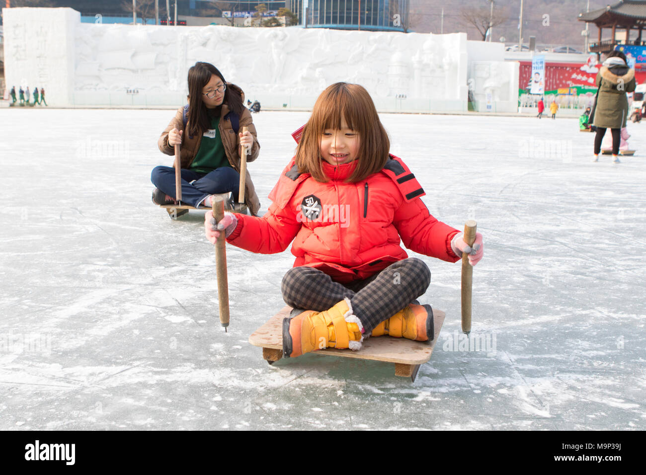 Zwei Mädchen sich nach vorn drücken, auf Schlitten an der Hwacheon Sancheoneo Ice Festival. Die Hwacheon Sancheoneo Ice Festival ist eine Tradition für die Menschen in Korea. Jedes Jahr im Januar Menschenmassen versammeln sich auf dem zugefrorenen Fluss der Kälte und dem Schnee des Winters zu feiern. Hauptattraktion ist Eisfischen. Jung und Alt warten geduldig auf ein kleines Loch im Eis für eine Forelle zu beißen. In zelten Sie können den Fisch vom Grill, nach dem sie gegessen werden. Unter anderem sind Rodeln und Eislaufen. Die in der Nähe Pyeongchang Region wird Gastgeber der Olympischen Winterspiele im Februar 2018. Stockfoto