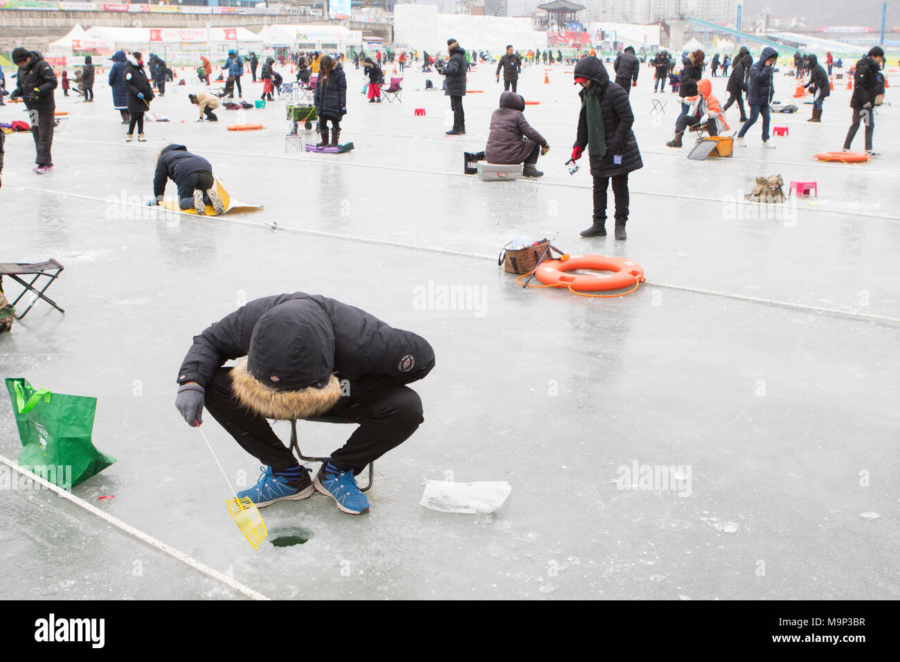 Menschen warten oben ein Loch in das Eis während der Eis angeln Festival an Hwacheon Sancheoneo in der Region Gangwon-do in Südkorea. Die Hwacheon Sancheoneo Ice Festival ist eine Tradition für die Menschen in Korea. Jedes Jahr im Januar Menschenmassen versammeln sich auf dem zugefrorenen Fluss der Kälte und dem Schnee des Winters zu feiern. Hauptattraktion ist Eisfischen. Jung und Alt warten geduldig auf ein kleines Loch im Eis für eine Forelle zu beißen. In zelten Sie können den Fisch vom Grill, nach dem sie gegessen werden. Unter anderem sind Rodeln und Eislaufen. Die in der Nähe Pyeongchang Region wird Gastgeber der Winter Stockfoto