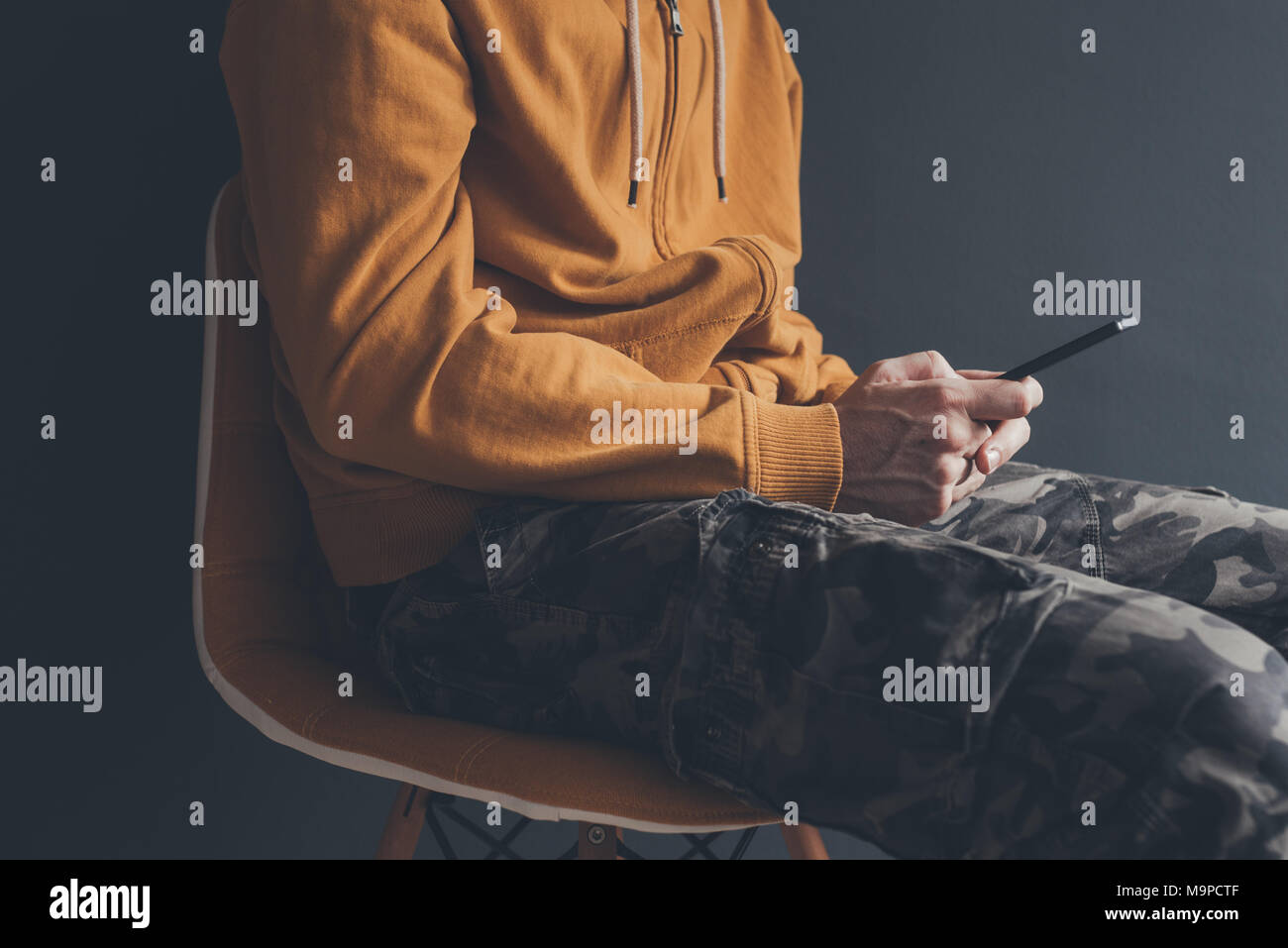 Legerer Mann mit smart phone laid back im Bürostuhl. Männliche Person tragen gelbe Hoodie und militärischen Hosen spielt mit seinem Handy. Stockfoto