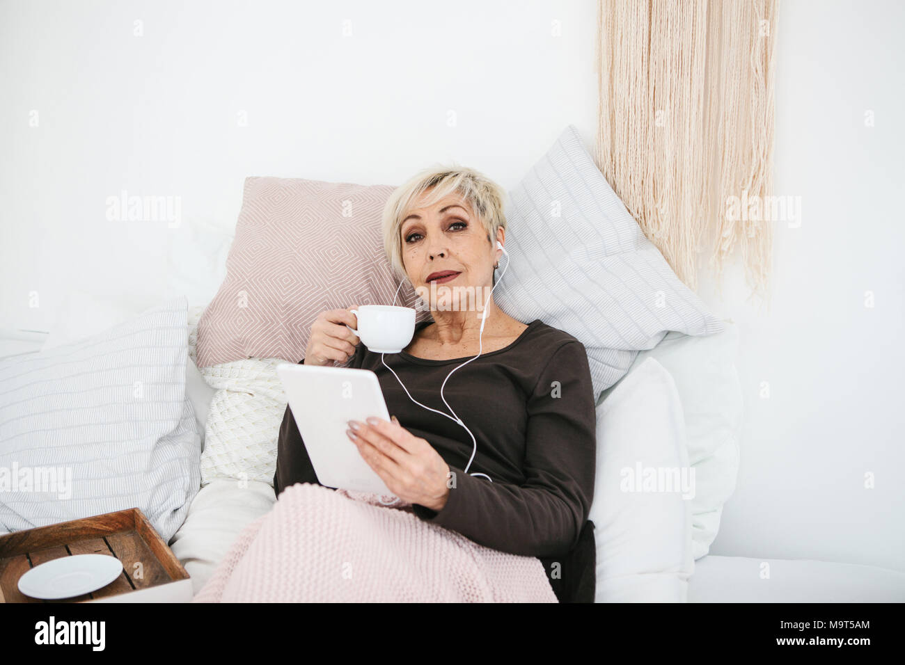 Eine positive Frau trinkt einen Kaffee am Morgen im Bett lag, und verwendet ein Tablet Videos ansehen, Musik hören und mit Freunden über soziale Netzwerke chat Stockfoto