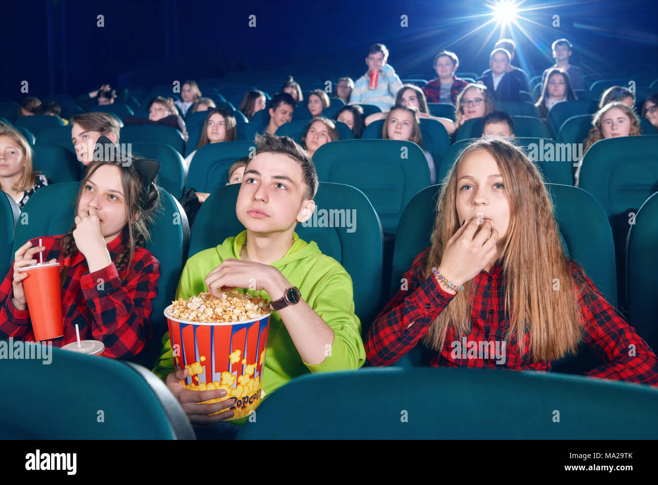 Drei Freunde essen Popcorn durch beobachten Film. Mädchen trinken kohlensäurehaltige Getränk von der großen roten können. Kinder beobachten neuen Film oder Cartoon. Suchen interessiert und verlassen. Tragen bunte Shirts. Stockfoto