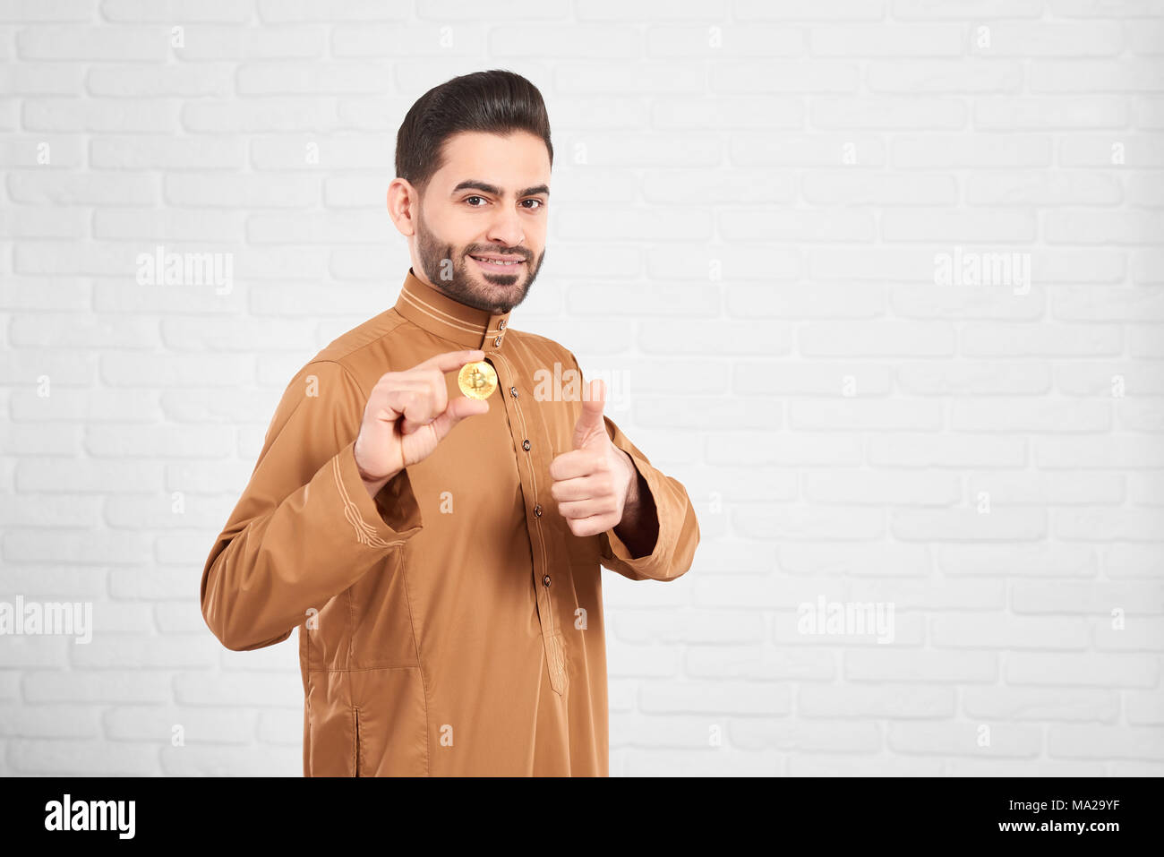 Junge attraktive muslimischer männlicher Modell in traditionelle islamische Kleidung, glücklich zu präsentieren goldene Bitcoin und Daumen hoch Vor unscharfen weiße Wand. Horizontale erschossen. Stockfoto