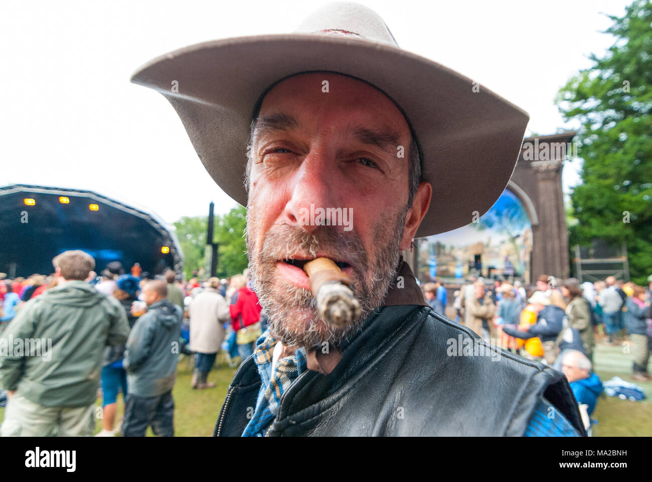 Bedeuten, bärtiger Mann als ein Cowboy Rauchen eine große Zigarre auf ein Musik Festival fancy dress Day gekleidet, Stockfoto