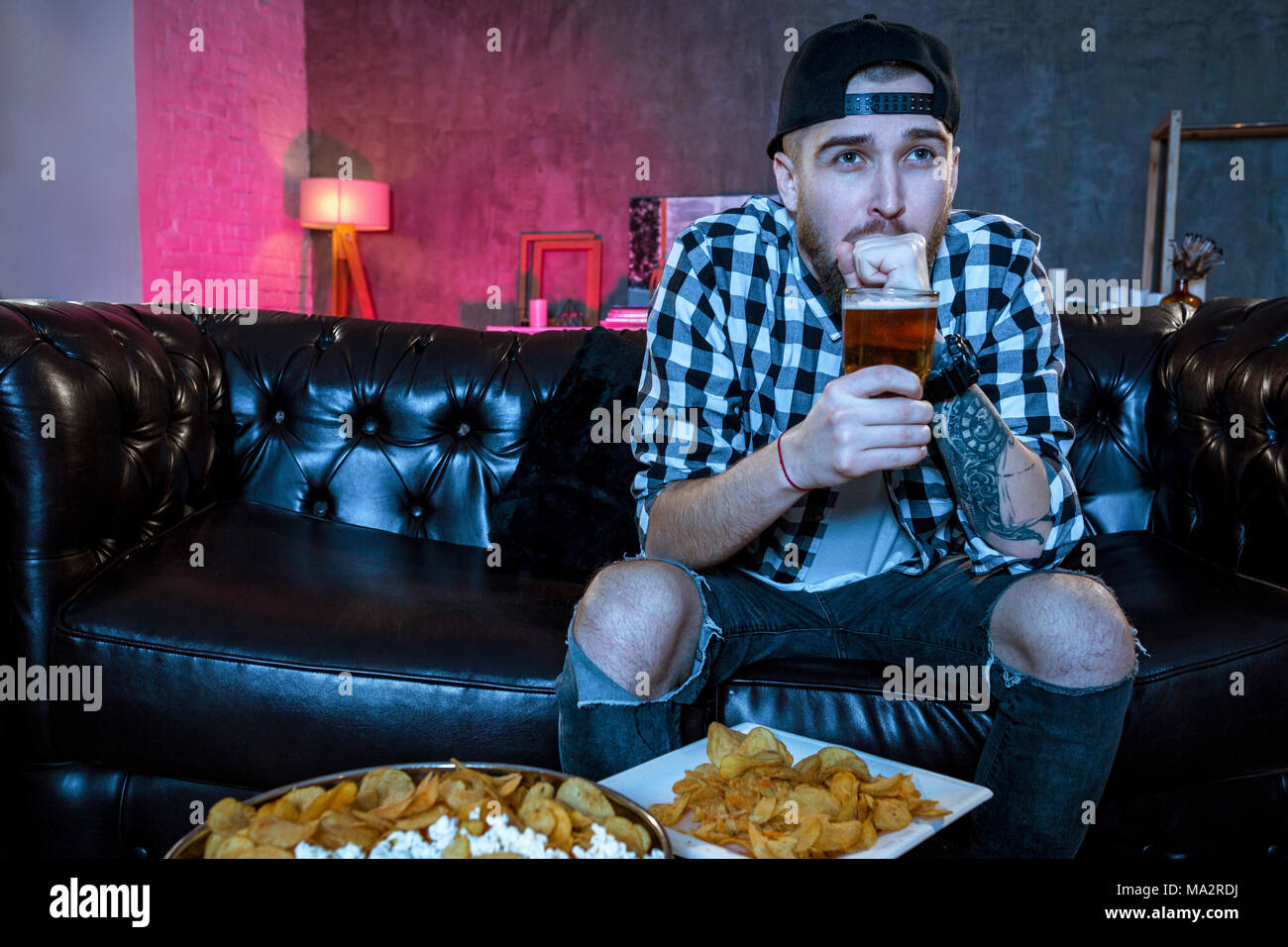 Junge fanatischer Mann, Fußball Spiel auf Fernsehen nervös und aufgeregt leiden Stress auf Sofa zu Hause mit Ball Bier Flasche und Chips aussehen Stockfoto