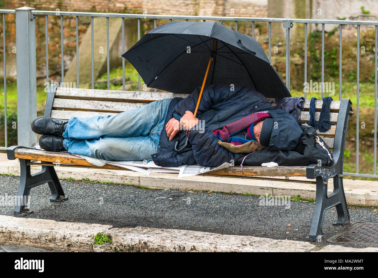 Ein obdachloser Mann im Regen auf einer Bank mit einem Regenschirm auf die Via dei Fori Imperiali historischen Zentrum von Rom, Latium, Italien. Stockfoto