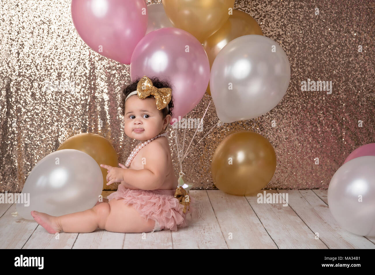 Ein Jahr alt, Baby Mädchen sitzen mit einem Bündel Luftballons. Sie trägt eine rosa blühende Pflanzen, Gold, pailletten Kopfbügel und eine Perlenkette. Stockfoto