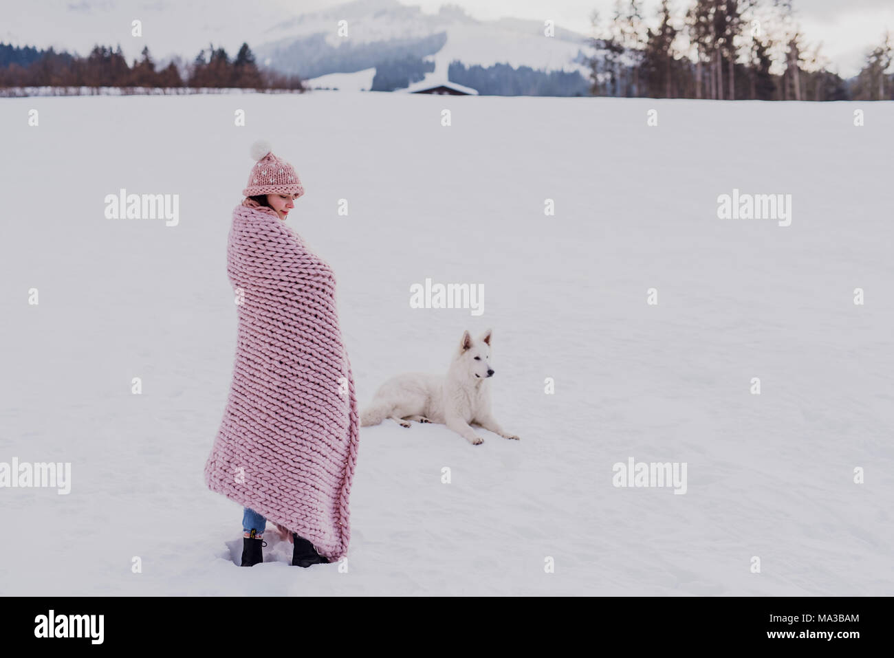 Junge Frau steht in einem rosa Decke in einer Winterlandschaft gewickelt, Hund liegt neben ihr im Schnee, Stockfoto