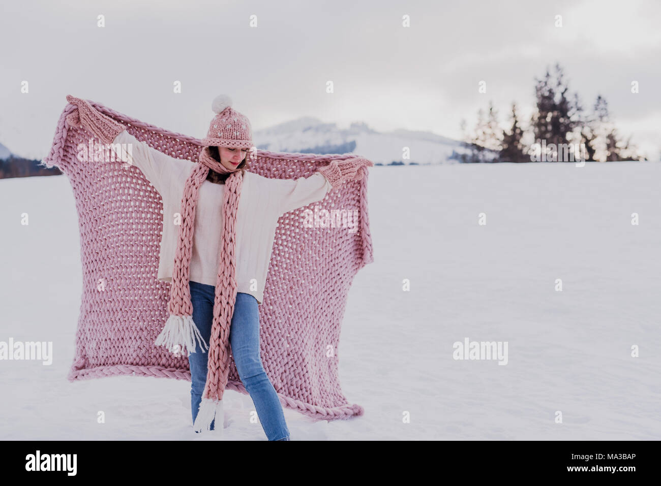 Junge Frau steht mit rosa Decke, wolle Mütze und Schal im Schnee, breitete die Arme, Stockfoto