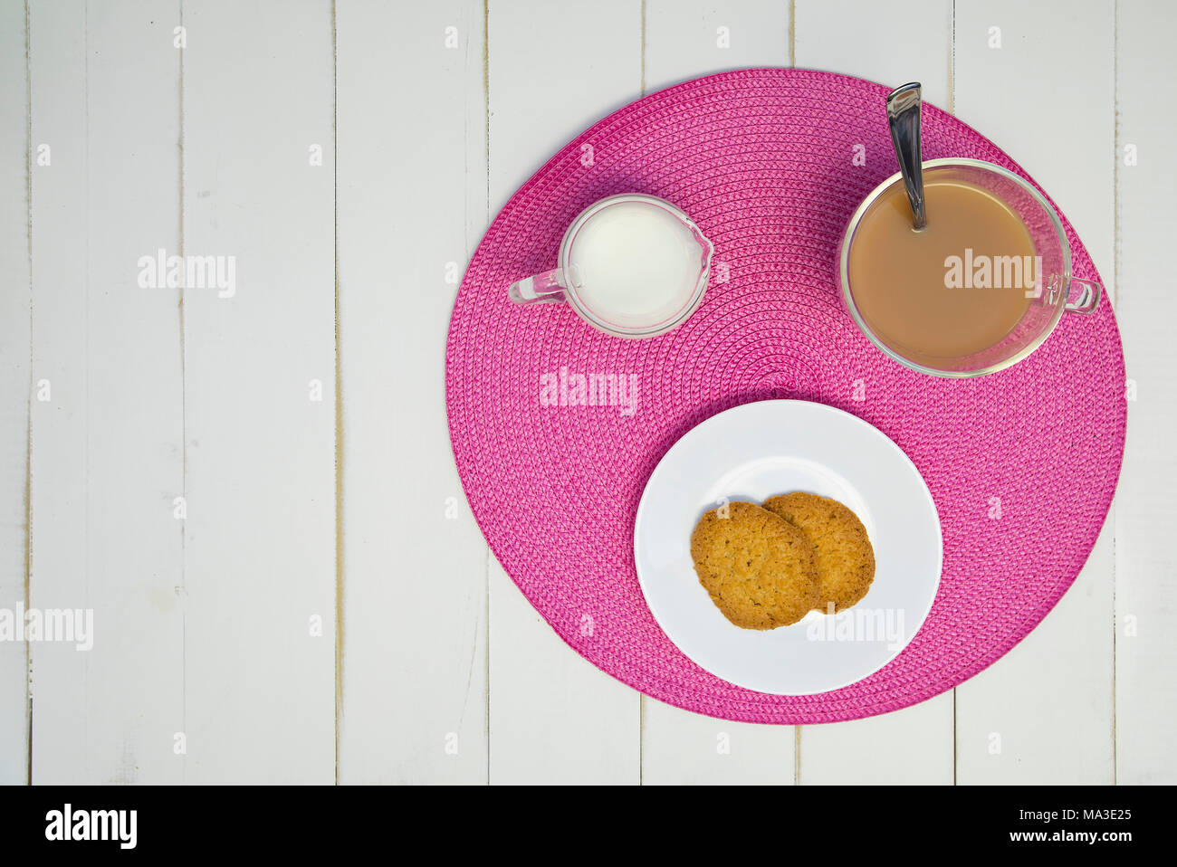 Heißen Tee, Kekse und Milch sind auf einem rosa Tischset auf eine weiße Tafel Tabelle angeordnet. Der Tee hat Milch in it. Die Anordnung ist außermittig und Schuß von Stockfoto