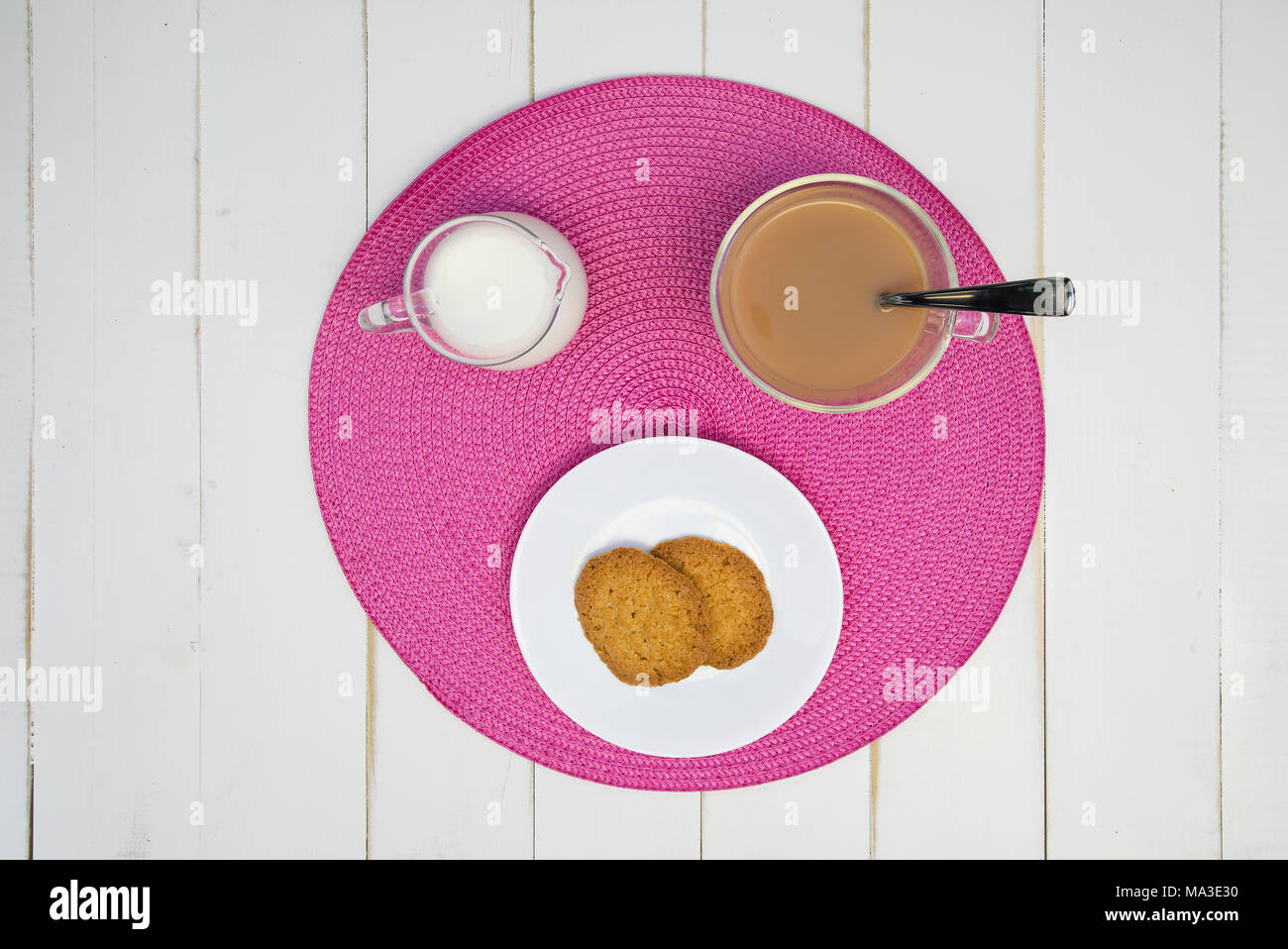 Heißen Tee, Kekse und Milch sind auf einem rosa Tischset auf eine weiße Tafel Tabelle angeordnet. Der Tee hat Milch in it. Die Anordnung wird zentriert und von o Shot Stockfoto