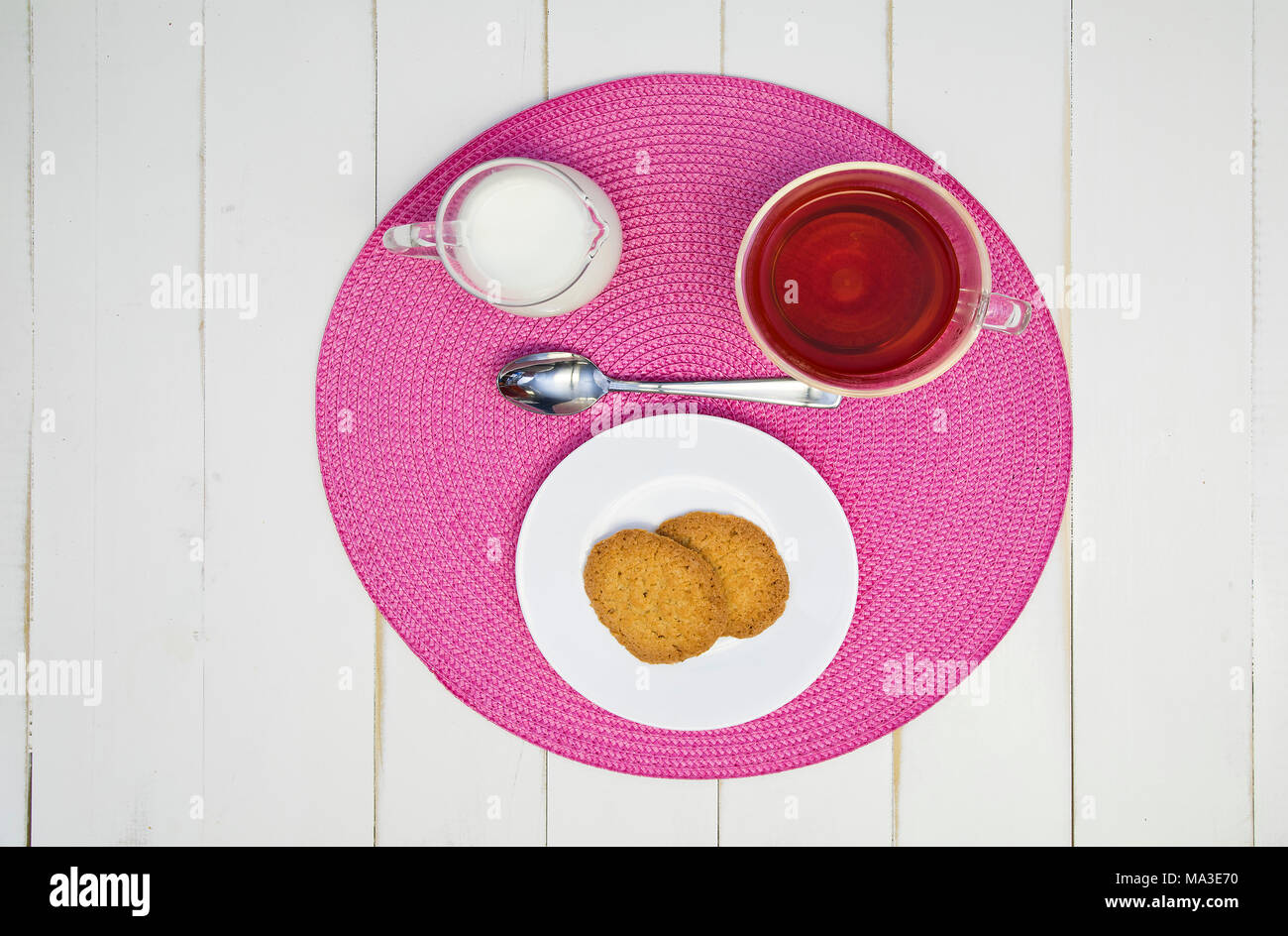 Heißen Tee, Kekse und Milch sind auf einem rosa Tischset auf eine weiße Tafel Tabelle angeordnet. Die Anordnung wird zentriert und von oben geschossen. Stockfoto