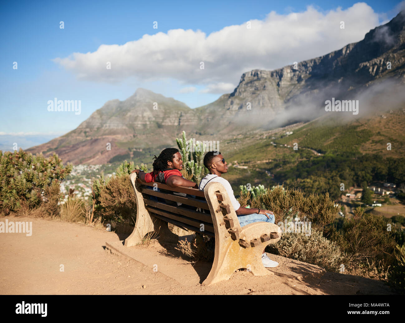 Zwei schwarze Jungs sitzen zusammen auf einer öffentlichen Bank neben einem Wanderweg mit Blick auf die bergige Gelände und mystische Wolken an einem warmen Su Stockfoto