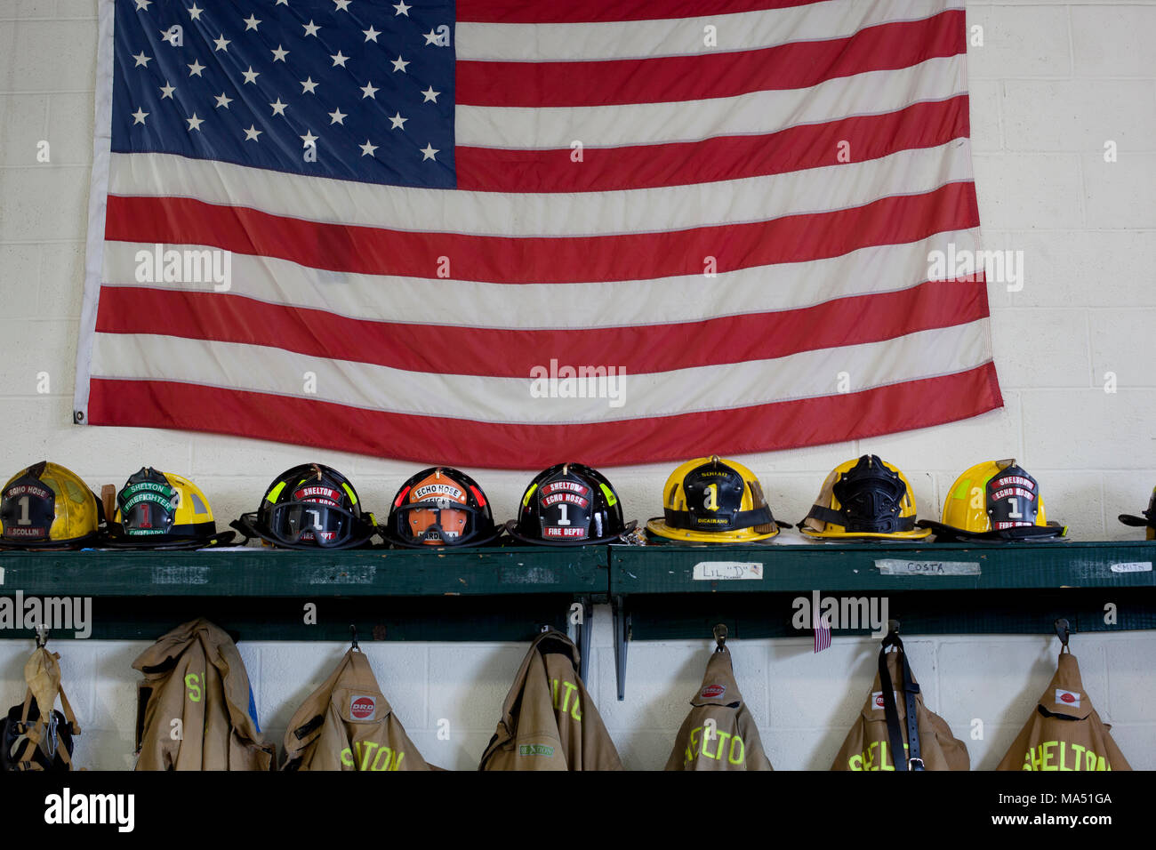 Schutzkleidung, Helme der Feuerwehrleute, und Wahlbeteiligung Mäntel zusammen mit einer amerikanischen Flagge auf Anzeige in der örtlichen freiwilligen Firehouse Stockfoto