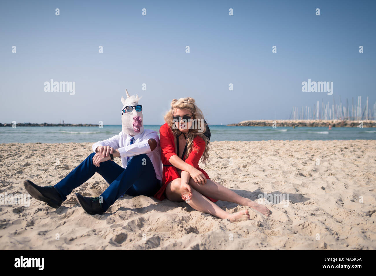 Ungewöhnliche Paar sitzt am Strand in der Nähe von Meer. Elegante Mann in lustige Maske und Sonnenbrille entspannt sich mit jungen Frau im roten Kleid. Freaky Einhorn in der Klage Stockfoto