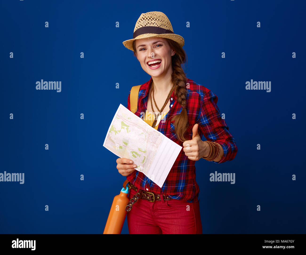 Auf der Suche nach inspirierenden Orten. glückliche junge Frau Wanderer mit Rucksack und Karte mit Daumen nach oben gegen den blauen Hintergrund Stockfoto