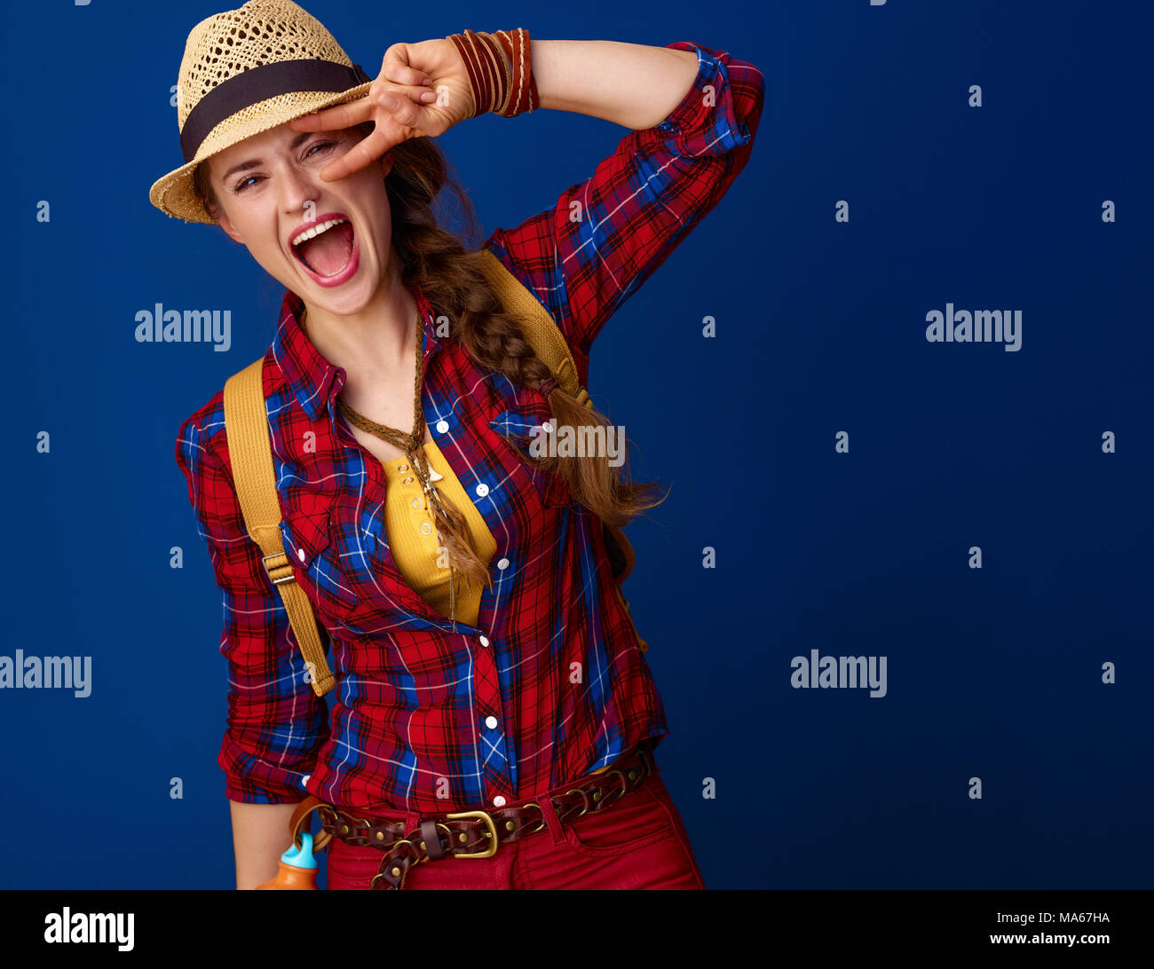 Auf der Suche nach inspirierenden Orten. glückliche junge Frau Wanderer in einen Plaid Shirt, Sieg Geste vor blauem Hintergrund Stockfoto