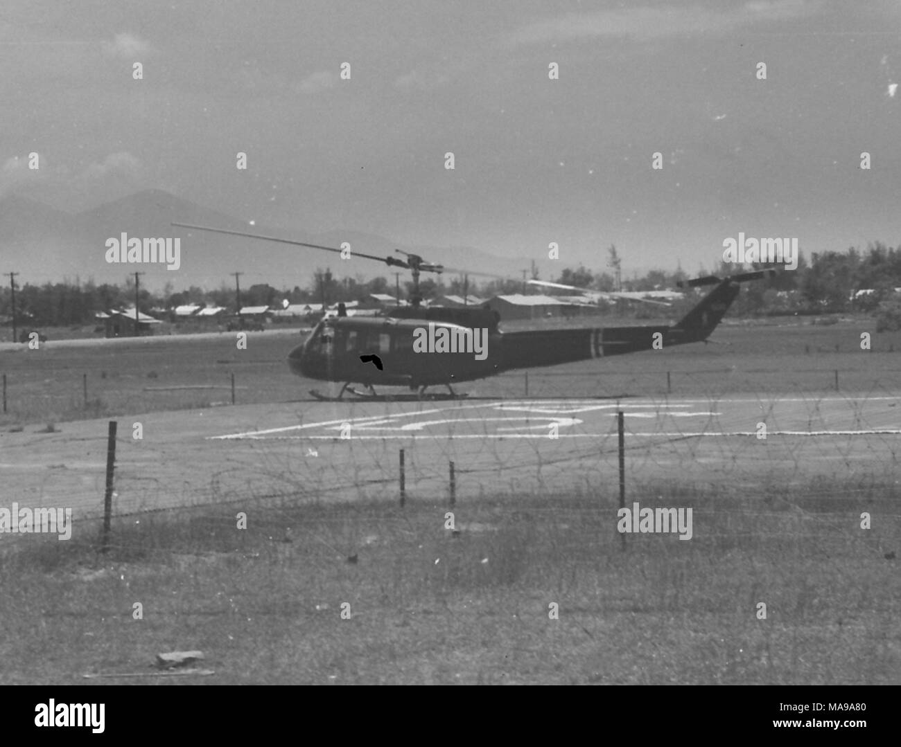 Schwarz-weiß Foto mit einem Hubschrauber auf einer Landebahn angedockt, mit Bäumen und Gebäuden, wahrscheinlich ein US Marine Corps Militärlager, in den Hintergrund, die in Vietnam während des Vietnam Krieges fotografiert (1955-1975), 1968. () Stockfoto