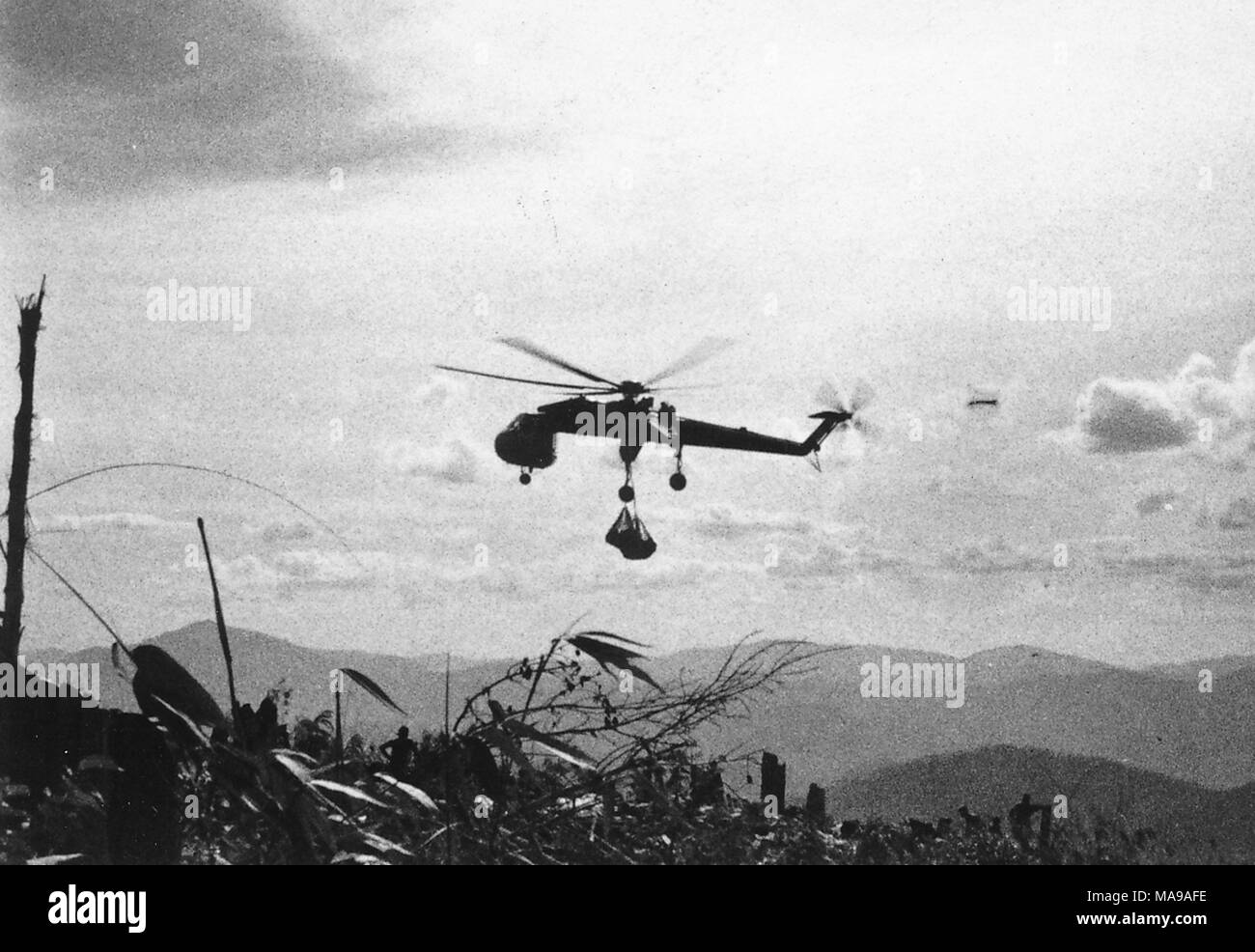 Schwarz-weiß Foto, ein Hubschrauber, mitten im Flug, große Taschen, und im Profil Silhouette gegen einen bewölkten Himmel, mit Bergen im Hintergrund, und Laub und ein Zaun im Vordergrund, die in Vietnam während des Vietnam Krieges fotografiert (1955-1975), 1971. () Stockfoto