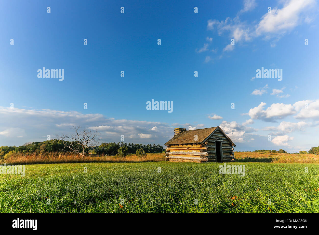 Eine einsame Blockhütte sitzt allein in der Mitte einer großen hellen, offenen Landschaft von Himmel und Felder bis an den Horizont an einem sommerlichen Nachmittag; mit Cop Stockfoto