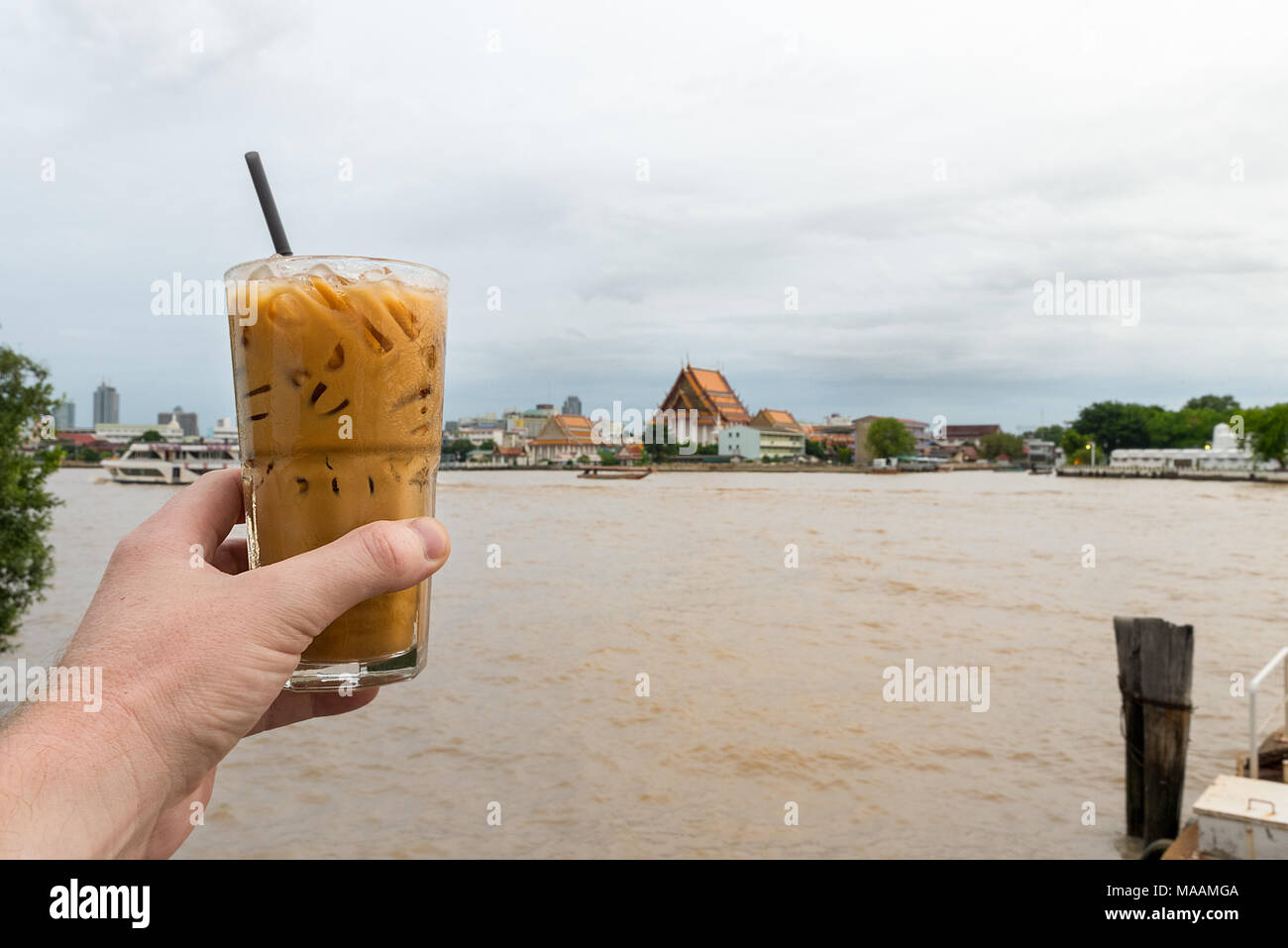 Eine männliche Hand hält ein klares Glas thai Eiskaffee mit Eiswürfeln und Kunststoff Stroh mit einem buddhistischen Tempel und großen Fluss im Hintergrund. Verbot Stockfoto
