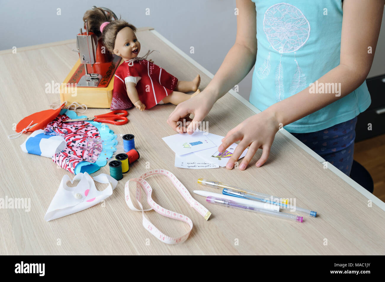 Die Hände der Mädchen halten die Skizzen der Kleidung für die Puppe. Auf dem Desktop gibt es Kugelschreiber, Bleistifte, Maßband, Stoff, Nähmaschine ein Stockfoto