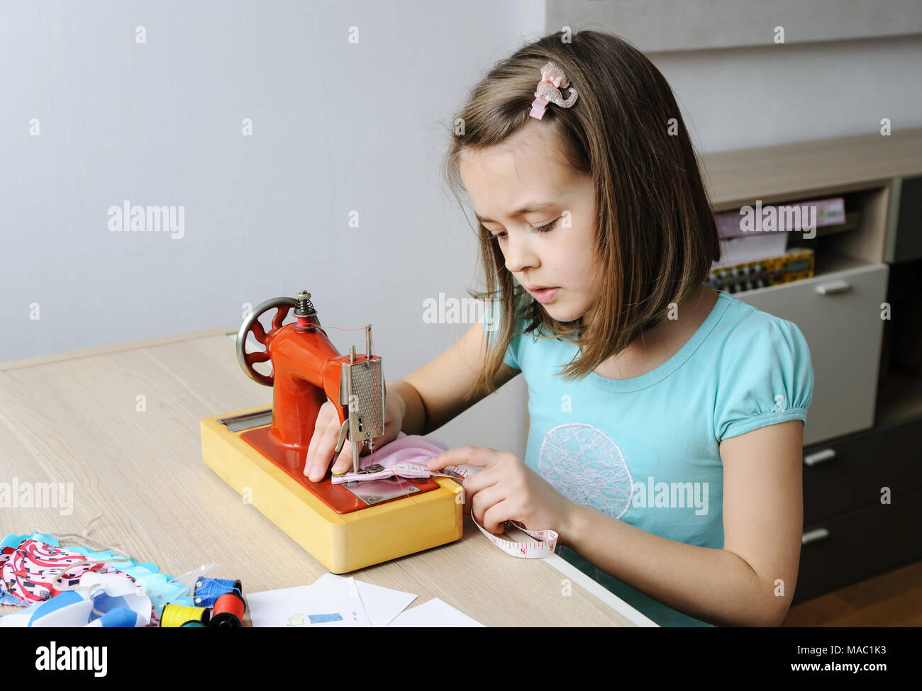 Das Mädchen ist das Nähen eines Kleides für eine Puppe auf einer Nähmaschine. Sie mit einem Maßband, das Produkt zu messen. Stockfoto