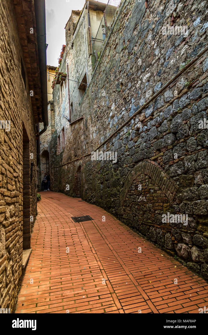 Oh, die ruhige Abgeschiedenheit und Entdeckung einer kleinen schattigen aus rotem Backstein, Spaziergang, von grossen steinernen Mauern und Gebäuden flankiert, Wicklung durch San Gimignano. Stockfoto