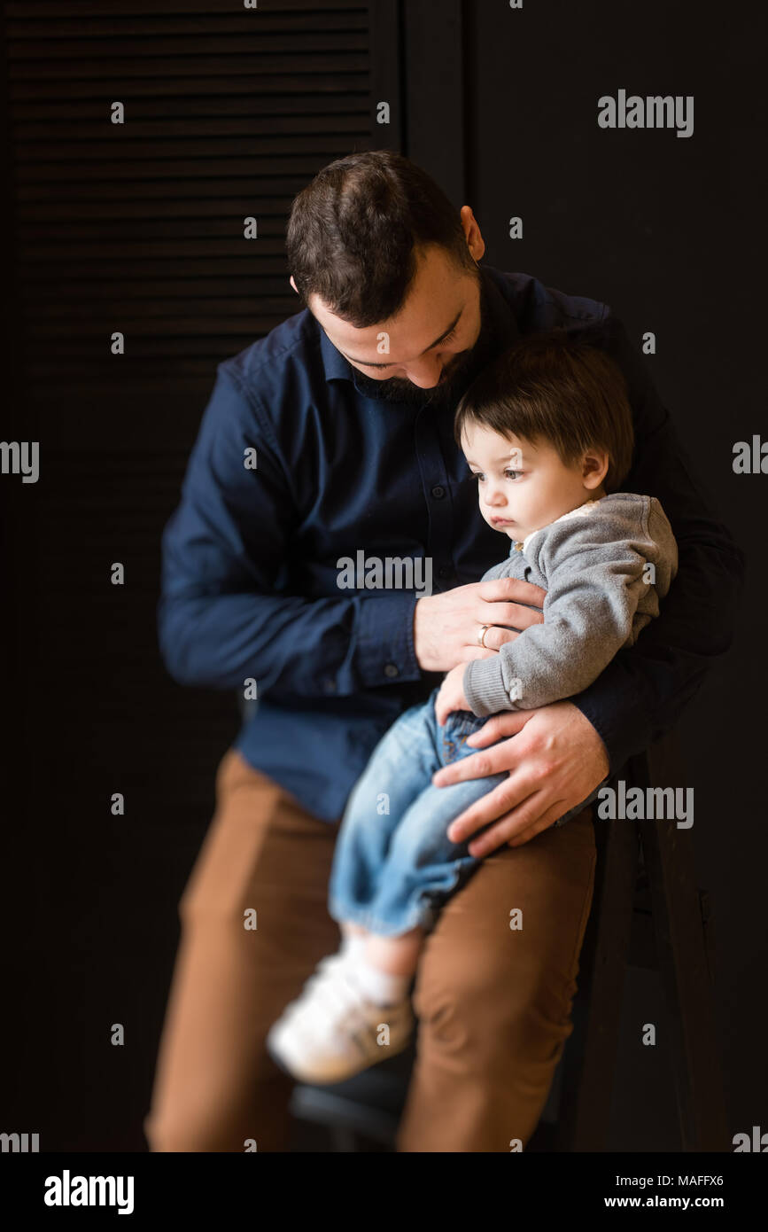 Kleines Baby sitzen in die Knie von Papa in Studio schwarzen Hintergrund. Family Portrait von Sohn und Vater. Stockfoto