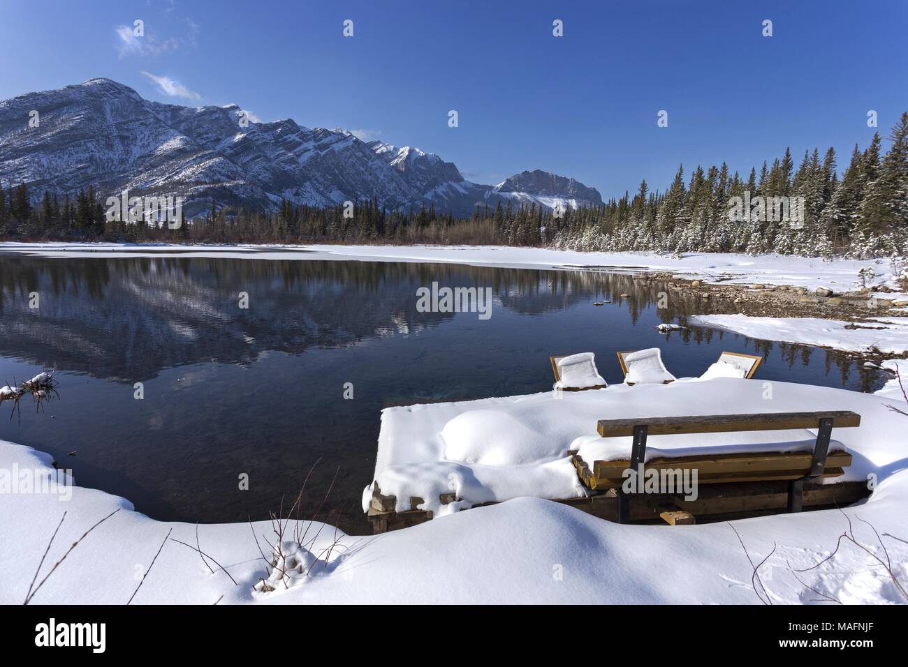 Parkbank bedeckt von Schnee durch ruhigen See, Anfang Frühling im Bow Valley Provincial Park, Alberta Ausläufern der kanadischen Rocky Mountains. Stockfoto