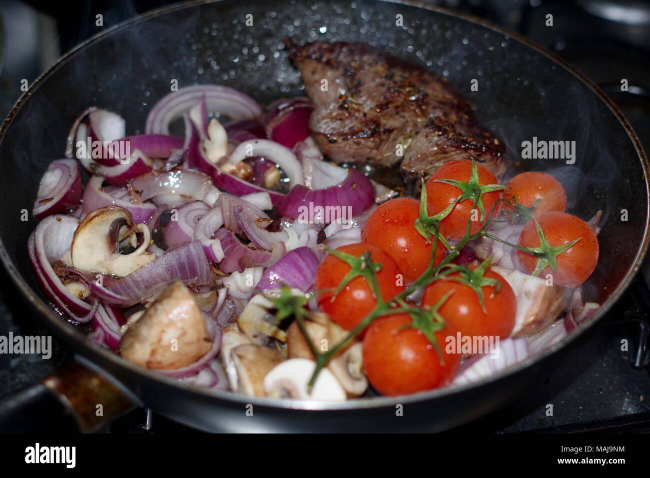 Steak zum Abendessen kochen in einer Pfanne, mit fröhlichen Tomaten, roten Zwiebeln und Pilze. Sauber, Essen und Kochen, UK. Stockfoto