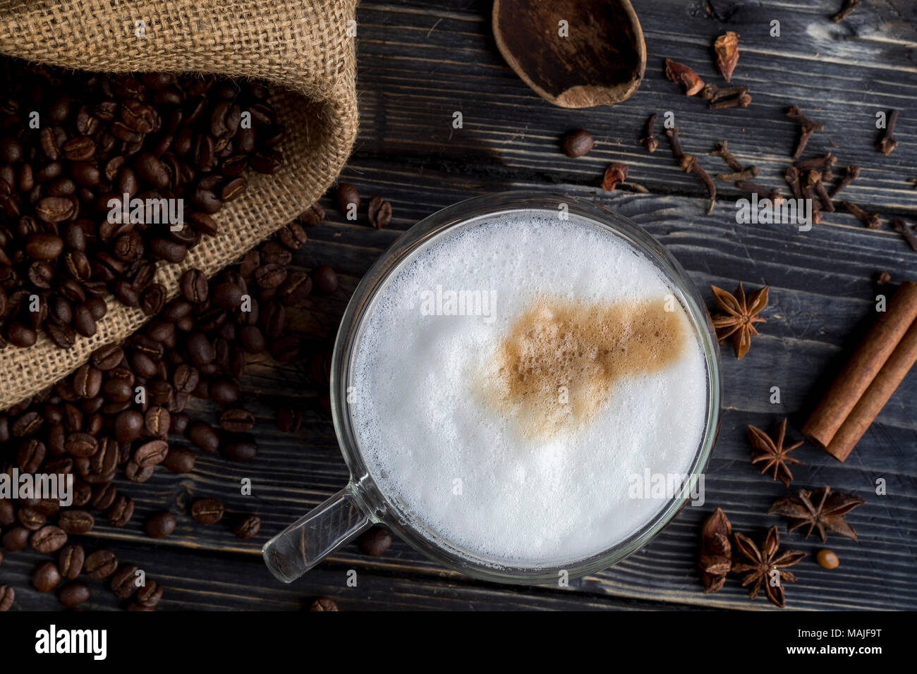 Tasse Cappuccino mit Schaum, Kaffeebohnen auf dem hölzernen Hintergrund, Jute Tasche und Gewürze Stockfoto