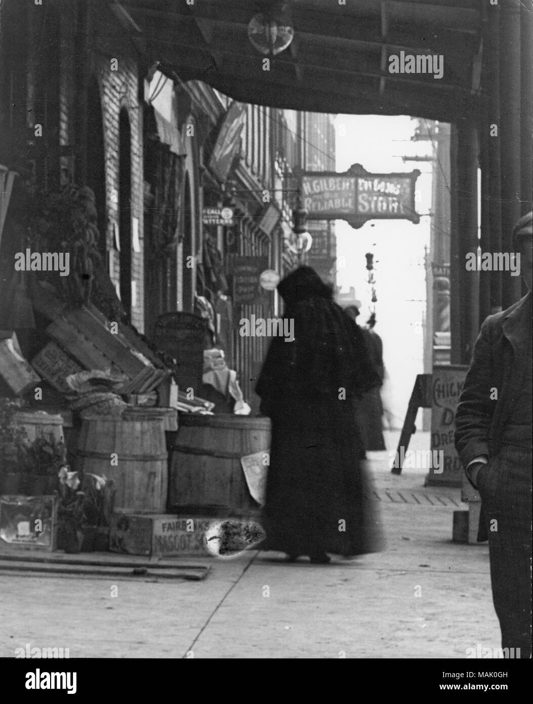 Vertikal, schwarz-weiß Foto einer Frau in Schwarz gekleidet von Kopf bis Fuß Durchlesen der Einkaufen geht. Im Hintergrund Zeichen für H. Gilbert speichern. 1909 Stadt Verzeichnis listen H. Gilbert trocken waren 2834 Manchester. Titel: Frau Einkaufen an einem fertigen Stall, vielleicht in der Nähe von 2834 Manchester. . Um 1910. Stockfoto