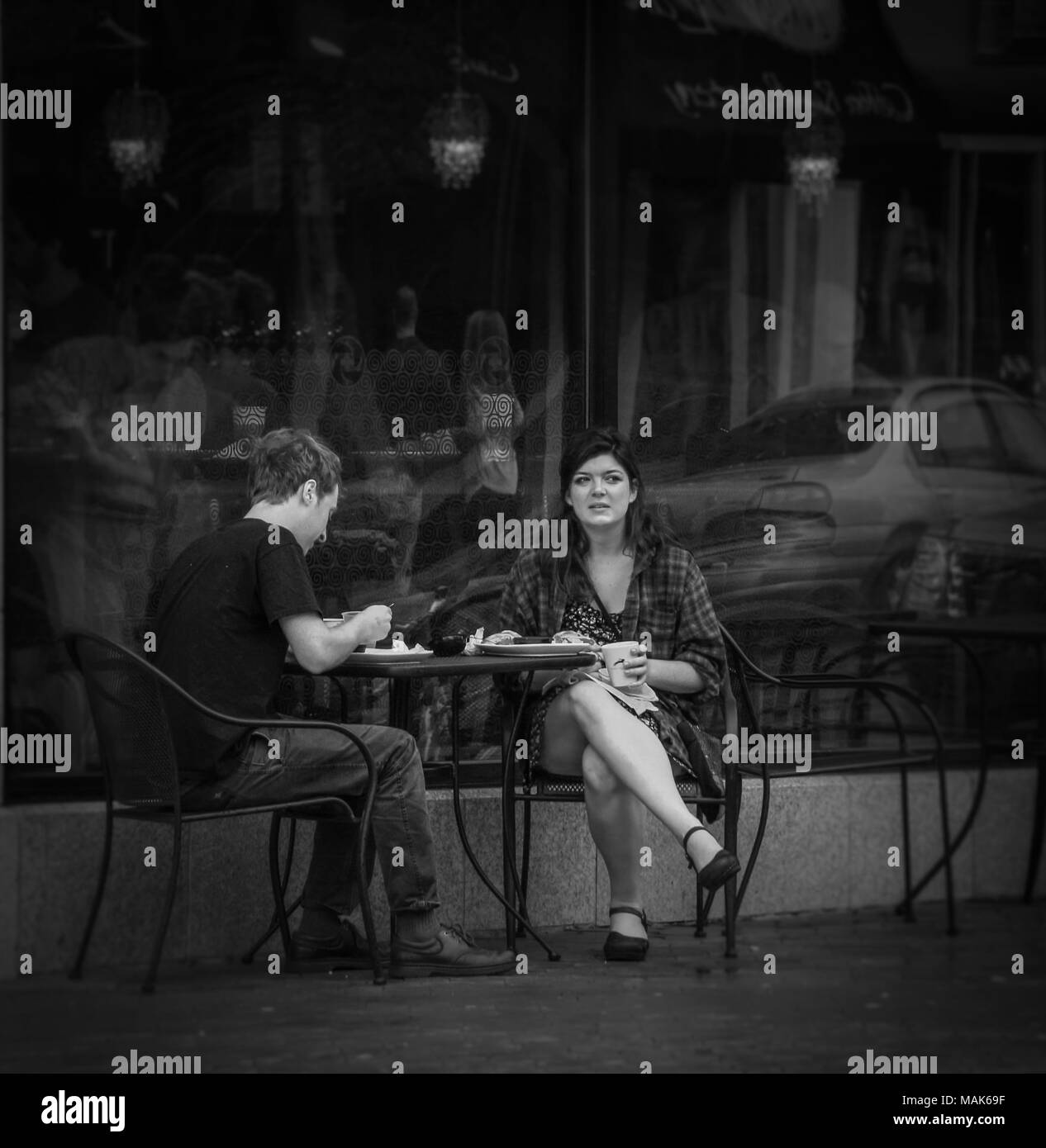 Junger Mann und Frau beim Mittagessen in einer Tabelle außerhalb Restaurant; vorbeifahrende Autos in das Glas hinter dem Paar widerspiegeln; die Frau nachdenklich schaut Stockfoto