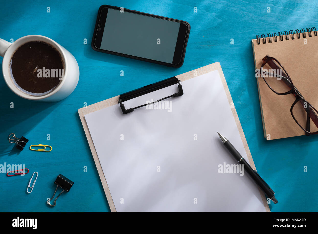 A4-Klemmbrett, Smartphone, kleines Notebook, Gläser, und Kaffee Tasse auf Blau Holz Tisch mit Natürlicher Lichteffekt in morgen Zeit. Hintergrund wit Stockfoto