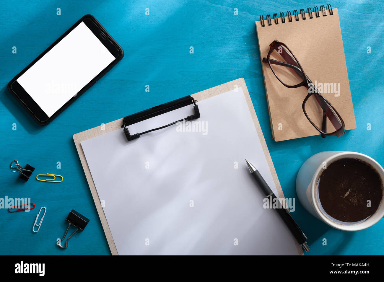 A4-Klemmbrett, Smartphone, kleines Notebook, Gläser, und Kaffee Tasse auf Blau Holz Tisch mit Natürlicher Lichteffekt in morgen Zeit. Hintergrund wit Stockfoto