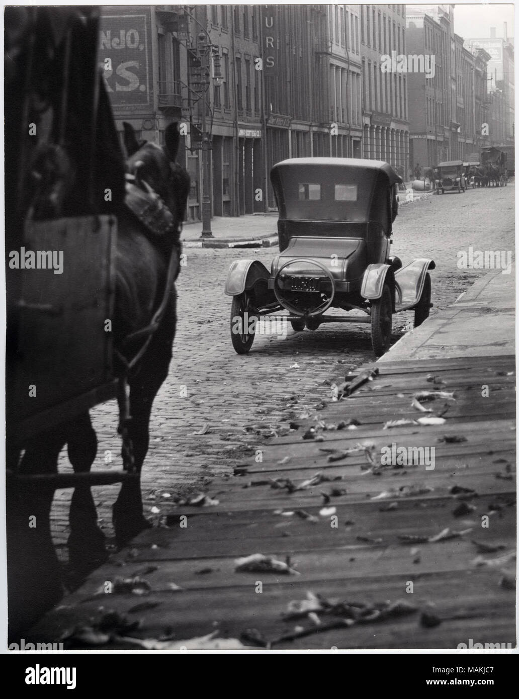 Vertikal, schwarz-weiß Foto zeigt eine 1910-Ära Auto an die Kandare eines nicht identifizierten gepflasterten Straße geparkt. Ein Pferd an einem Wagen oder Wagen kann auf der linken Seite des Bildes gesehen werden, und andere Autos und Wagen sind weitere sichtbare auf der Straße. Gebäude an der gegenüberliegenden Seite der Straße, obwohl die Zeichen sind schwer zu lesen. Eine hölzerne Gehweg kann in der unteren rechten Ecke gesehen werden. Titel: Auto am Strassenrand eines nicht identifizierten gepflasterten Straße geparkt. . Um 1910. Holt, Charles Clement, 1866-1925 Stockfoto