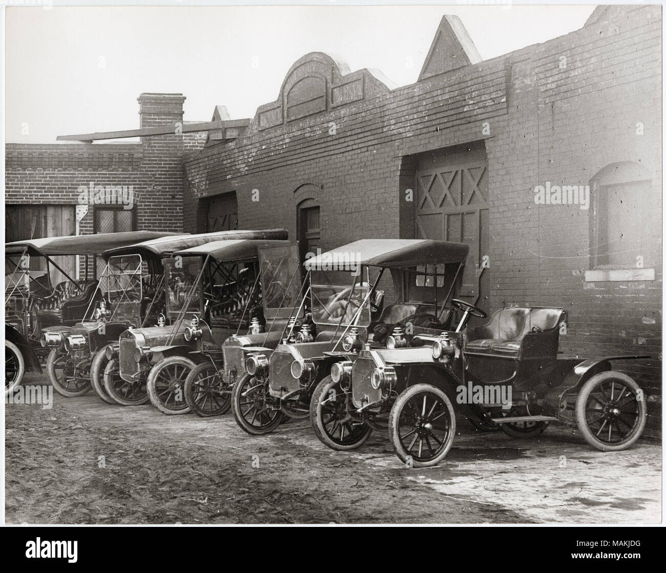 Horizontal, schwarz-weiß Foto zeigt eine Linie der 1910-Ära Automobile vor der alten Stadt Garage geparkt. Einige der Wagen geöffnet sind, während andere mit einem Dach abgedeckt sind. Die Garage ist ein L-förmiges, 1-stöckiges Ziegelgebäude. Titel: Linie 1910 - ära Automobile vor der alten Stadt Garage geparkt. . Um 1910. Holt, Charles Clement, 1866-1925 Stockfoto