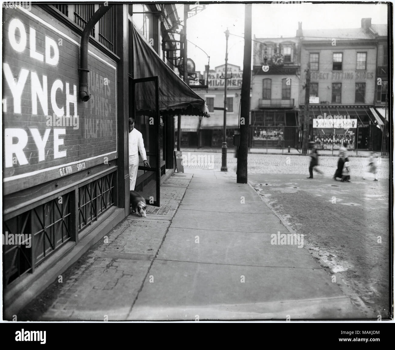 Horizontal, schwarz-weiß Foto, das einen Mann zeigt, dass ein Hund durch die Hintertür einer Limousine in 1151 South Broadway. Der Mann in Weiß gekleidet, trägt einen weißen Kittel oder Schürze. Ein Schild an der Seite der Limousine lackiert wirbt für alte Lynch Roggen. Mehrere Unscharfe Figuren, darunter eine Frau und Kinder, kann man über die Straße auf der rechten Seite des Fotos, wahrscheinlich Hickory Street. Anzeichen für eine Hutmacherin und Schuhgeschäft kann über South Broadway gesehen werden. Titel: der Mann, der einen Hund aus der Hintertür einer Limousine in 1151 South Broadway. . Ca. 1907. Stockfoto